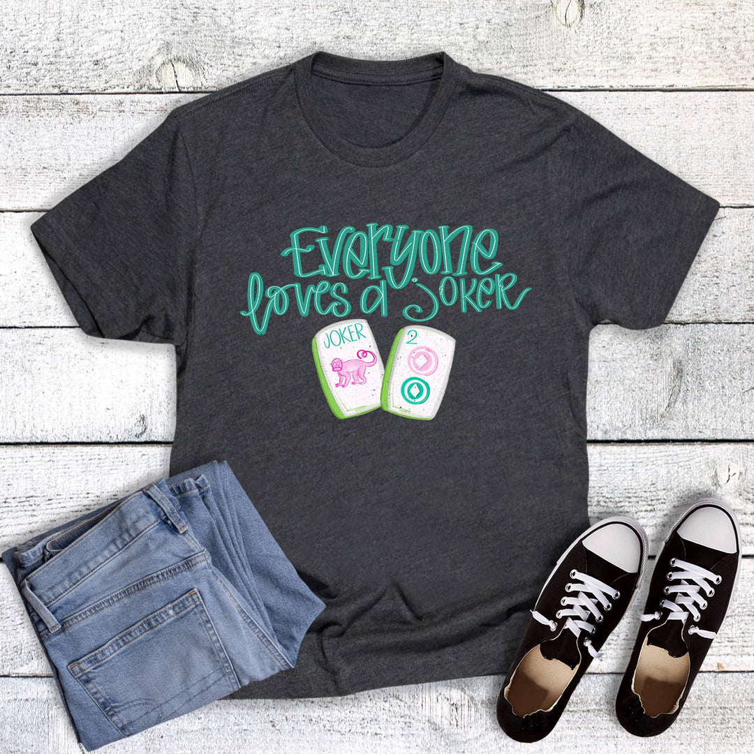 Charcoal gray graphic T-shirt featuring the phrase “Everyone Loves a Joker” with colorful mahjong tiles, styled with light-wash jeans and black sneakers.